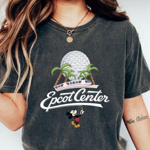 May include: Dark gray t-shirt featuring a graphic of the Epcot Center logo with palm trees, a monorail, and Mickey Mouse. The text "Epcot Center" is in a white script font. The shirt has a vintage, worn look.