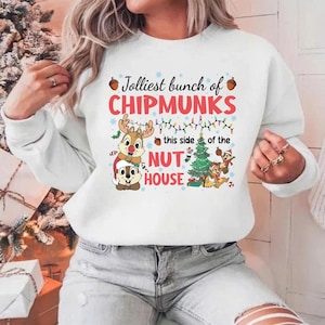 Peut inclure: Sweat-shirt blanc avec un motif coloré de Chip et Dale en tenues de Noël, avec le texte "Jolliest bunch of CHIPMUNKS" et "Nut House". Le motif comprend un sapin de Noël et des décorations festives.