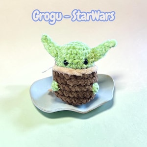 Peut inclure: Une petite figurine en crochet faite à la main représentant une créature verte aux grandes oreilles, vêtue d'un vêtement marron texturé en forme de nacelle. Elle a des yeux en perles noires et repose sur un plat en céramique bleu clair. Texte : Grogu-StarWars.