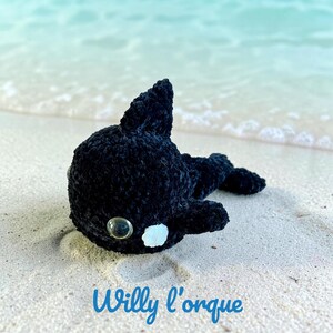 Peut inclure: Une peluche orque crochetée noire avec un corps arrondi, une nageoire dorsale et des nageoires latérales. Le jouet présente deux grands yeux de sécurité en plastique transparent et une petite tache circulaire blanche près de l'œil. Le texte "Willy l'orque" est imprimé en bleu.
