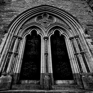 May include: A grayscale image of a cathedral entrance. The arched doorway features ornate carvings and dark, detailed doors. Above, a series of arched windows and stone brickwork complete the architectural composition. The image is taken from a low angle.