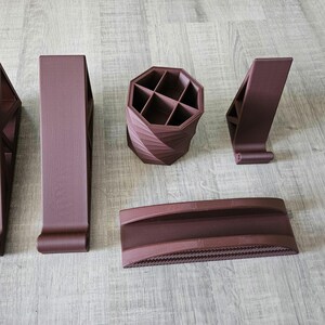 Peut inclure: Ensemble d'organisateurs de bureau marron imprimés en 3D. L'ensemble comprend deux supports angulaires, un porte-stylos à compartiments, un support de téléphone et un long plateau à base texturée. Les articles sont disposés sur une surface imitation bois clair.