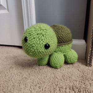May include: A handmade crochet turtle in shades of green. The turtle has a round head, a darker green shell, and small black eyes. The turtle is sitting on a beige carpet.