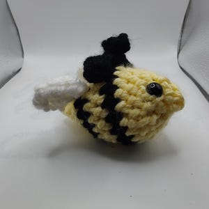 May include: A handmade crochet bee with yellow and black stripes, black antennae, and white wings. The bee has a small black bead for an eye. The craft item is sitting on a white surface.