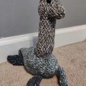 May include: A handmade knitted Nessie monster toy in shades of gray and blue. The Loch Ness monster plush has a long neck, small head, and flipper-like appendages. The toy is made of soft yarn and has a cute, friendly appearance.