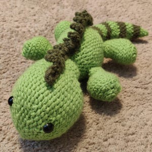 May include: A handmade crochet lizard toy in shades of green and brown. The lizard features a textured body, small black eyes, and a detailed spine. The tail has brown stripes. This soft toy is suitable for children.