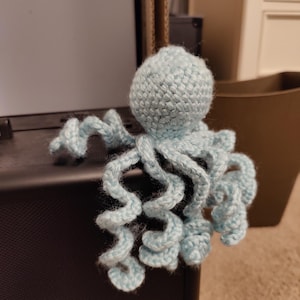 May include: A light blue crocheted octopus plush toy featuring a round head and eight long, spiraled tentacles. The yarn texture is visible throughout the entire structure of the handmade creature, which is crafted from soft, light blue yarn.
