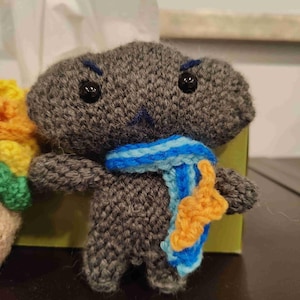 Może przedstawiać: Ręcznie robiona szara pluszowa zabawka z czarnymi guzikami i niebiesko-pomarańczowym szalikiem. Zabawka ma małe ramiona i nogi oraz wyszywane niebieskie usta i brwi. Tło to pudełko po chusteczkach.