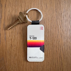 Peut inclure: Un porte-clés rectangulaire blanc avec un design de cassette VHS rétro, avec le texte "Dynamic T-120" et une bande colorée. Une clé dorée est attachée à l'anneau du porte-clés, posée sur une surface en bois.