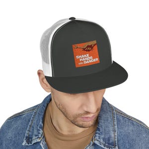Op de afbeelding: Zwarte en witte truckerpet met een platte klep. Het voorpaneel is donkergrijs met een oranje vierkante afbeelding met de tekst "SHAKE HANDS with DANGER". De afbeelding toont een hand die een moersleutel vasthoudt.