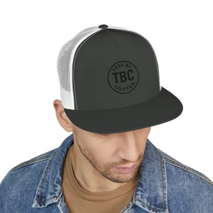 May include: A dark gray trucker hat with a white mesh back and a black brim. The hat features a circular logo with the text "TEES BY TBC COTTER". The hat is worn by a person wearing a denim jacket.