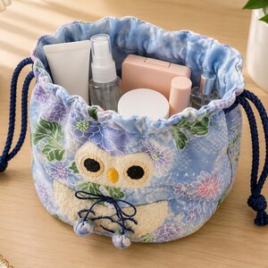 Peut inclure: Un sac à cosmétiques bleu à cordon de serrage avec un motif floral et hibou. Le sac contient divers produits cosmétiques. Il est doté d'une fermeture à cordon bleue et est posé sur une surface en bois.