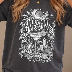 May include: A charcoal grey short-sleeve t-shirt featuring a white graphic print with the text "MIDNIGHT MARGARITAS". The design includes a margarita glass, a crescent moon, a frog, candles, books, and floral elements in a detailed illustration.