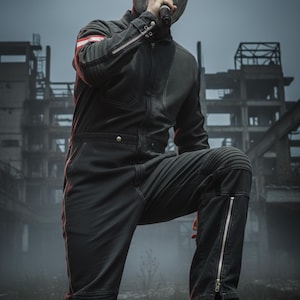 Könnte beinhalten: Eine Person in einem schwarzen Overall mit einem rot-weißen Streifen am Arm, der ein Mikrofon hält. Der Overall hat Reißverschlüsse an den Beinen und befindet sich vor dem Hintergrund eines baufälligen Gebäudes.