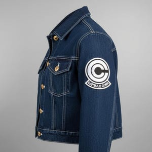 Puede incluir: Chaqueta vaquera azul oscuro con costuras blancas, botones dorados y un parche de Capsule Corp en la manga. La chaqueta tiene dos bolsillos en el pecho y un cuello clásico. Una adición elegante a cualquier guardarropa.