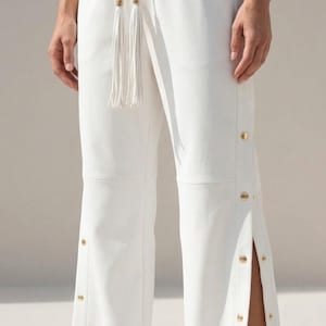 May include: White drawstring pants with gold-colored button details and side slits. The pants feature a drawstring waist with tassel accents. The model is wearing white sneakers with gold-colored toes.