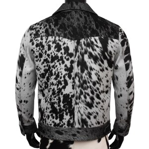 K&ouml;nnte beinhalten: Kuhfelljacke mit einem schwarz-wei&szlig; gefleckten Muster. Die Jacke hat einen schwarzen Kragen und Besatz, und die R&uuml;ckseite ist sichtbar. Das gefleckte Muster ist eine Mischung aus Schwarz und Wei&szlig; und sorgt f&uuml;r einen einzigartigen und stilvollen Look.