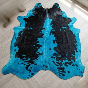Op de afbeelding: Een koeienvacht tapijt met een opvallend design. Het tapijt heeft een levendige turquoise basis met grote, onregelmatige zwarte vlekken. De vorm van het tapijt volgt de natuurlijke omtrek van de huid, wat bijdraagt aan de unieke esthetiek. Dit tapijt is een decoratieve vloerbedekking.