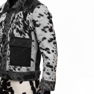 K&ouml;nnte beinhalten: Schwarz-wei&szlig;e Kuhfelljacke mit Knopfleiste, schwarzer Tasche und Kragen. Die Jacke hat ein gepunktetes Muster und ein klassisches Design.