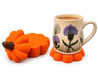 Puede incluir: Una taza de cerámica con esmalte color crema y flores de cardo moradas pintadas sobre un posavasos naranja con forma de flor. Al lado, un segundo posavasos naranja tiene encima una tapa naranja con forma de calabaza.
