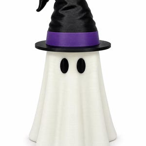 Puede incluir: Una figura de fantasma blanca con un cuerpo liso y drapeado y dos ojos ovalados negros. El fantasma lleva un sombrero de bruja negro puntiagudo con una banda morada ancha en la base. El objeto est&aacute; de pie sobre un fondo blanco liso.