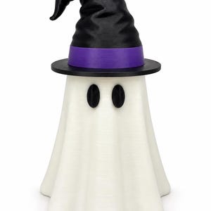 Puede incluir: Una figura de fantasma blanco que lleva un sombrero de bruja negro con una banda morada. El fantasma tiene dos ojos negros ovalados y un cuerpo drapeado y fluido. El sombrero presenta una parte superior puntiaguda y un ala ancha y negra.