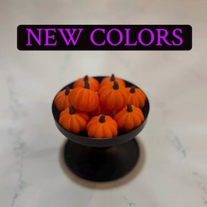 May include: A black pedestal bowl filled with small, bright orange pumpkin-shaped objects, each featuring a dark brown stem. Above the bowl, a black rectangular banner displays the text NEW COLORS in bold, bright purple capital letters.