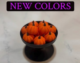 Puede incluir: Un cuenco negro con pedestal lleno de pequeños objetos de color naranja brillante con forma de calabaza, cada uno con un tallo marrón oscuro. Sobre el cuenco, una pancarta rectangular negra muestra el texto NEW COLORS en letras moradas.