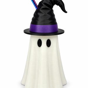 Puede incluir: Una figura de fantasma blanco que lleva un sombrero de bruja negro con una banda morada. El fantasma tiene dos ojos ovalados negros y una pajita de colores del arco&iacute;ris que sobresale de la parte superior del sombrero con textura vertical.