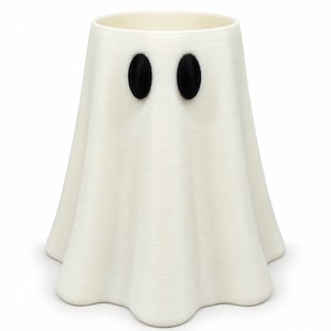 Puede incluir: Un recipiente cil&iacute;ndrico y hueco de color blanco con forma de fantasma y una base acampanada y ondulada. El objeto presenta dos ojos negros ovalados y prominentes, orientados verticalmente, situados cerca de la parte superior.