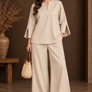 Peut inclure: Un ensemble deux pièces beige composé d'un haut à manches longues avec une encolure fendue et des poignets ouverts, assorti d'un pantalon large. La tenue est complétée par un sac à main en paille tressée et des sandales ouvertes beiges.