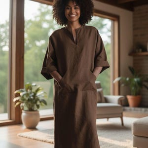 May include: A woman wearing a brown linen caftan dress with a V-neck and pockets. The dress has elbow-length sleeves and a relaxed fit. The woman is smiling in a well-lit room with large windows and plants.