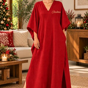 May include: A floor-length red kaftan dress featuring a V-neckline, wide short sleeves, and side slits. The fabric is solid red with the word Christmas embroidered in gold cursive script on the upper left chest area of the garment.