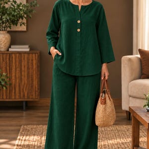 Op de afbeelding: Een donkergroene tweedelige set bestaande uit een tuniek met lange mouwen, een split-halslijn en twee lichte knopen, gecombineerd met een bijpassende broek met wijde pijpen. De outfit wordt gedragen met beige slippers en een gevlochten rieten tas.