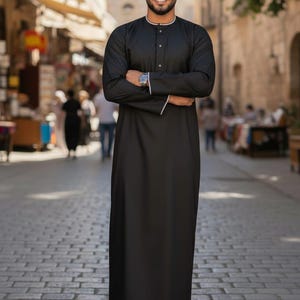 Op de afbeelding: Een zwarte thobe, een traditioneel Arabisch kledingstuk, met witte bies op de kraag en manchetten. De lange mouwen jurk wordt gedragen met bruine sandalen. De persoon staat op een geplaveide straat.