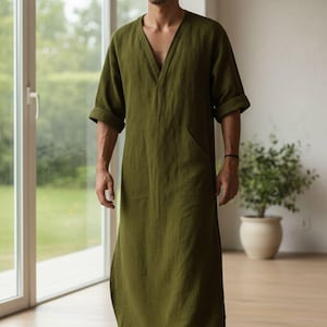 May include: Olive green linen robe with a V-neck and short sleeves. The robe has two front pockets and a floor-length hem. The model is standing in front of a large window with a plant in the background.