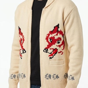 Puede incluir: Una chaqueta de punto beige con cremallera, cuello chal y dos bolsillos delanteros. La prenda presenta un gráfico de cabeza de zorro rojo y negro a cada lado del pecho y un pequeño borde con patrón gris a lo largo del dobladillo y los puños.