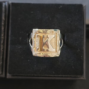 Op de afbeelding: Een goudkleurige zegelring met een vierkante voorkant met de letter "K". De letter is zilverkleurig, tegen een gouden achtergrond. De ring wordt gepresenteerd op een zwarte ondergrond.