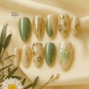 May include: A collection of press-on nails in various designs. Some nails are solid sage green or cream, while others feature gold leaf accents, floral designs, and a 3D flower. The brand name "Press & Blink" is visible in the upper left corner.
