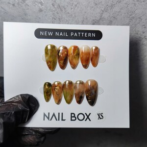 以下が含まれることがあります： Nail Box XS パッケージに入ったアーモンド型のつけ爪のセット。爪は、茶色、金、緑の色合いの大理石模様で、金箔のアクセントが付いています。パッケージには「NEW NAIL PATTERN」と「NAIL BOX」の文字があります。