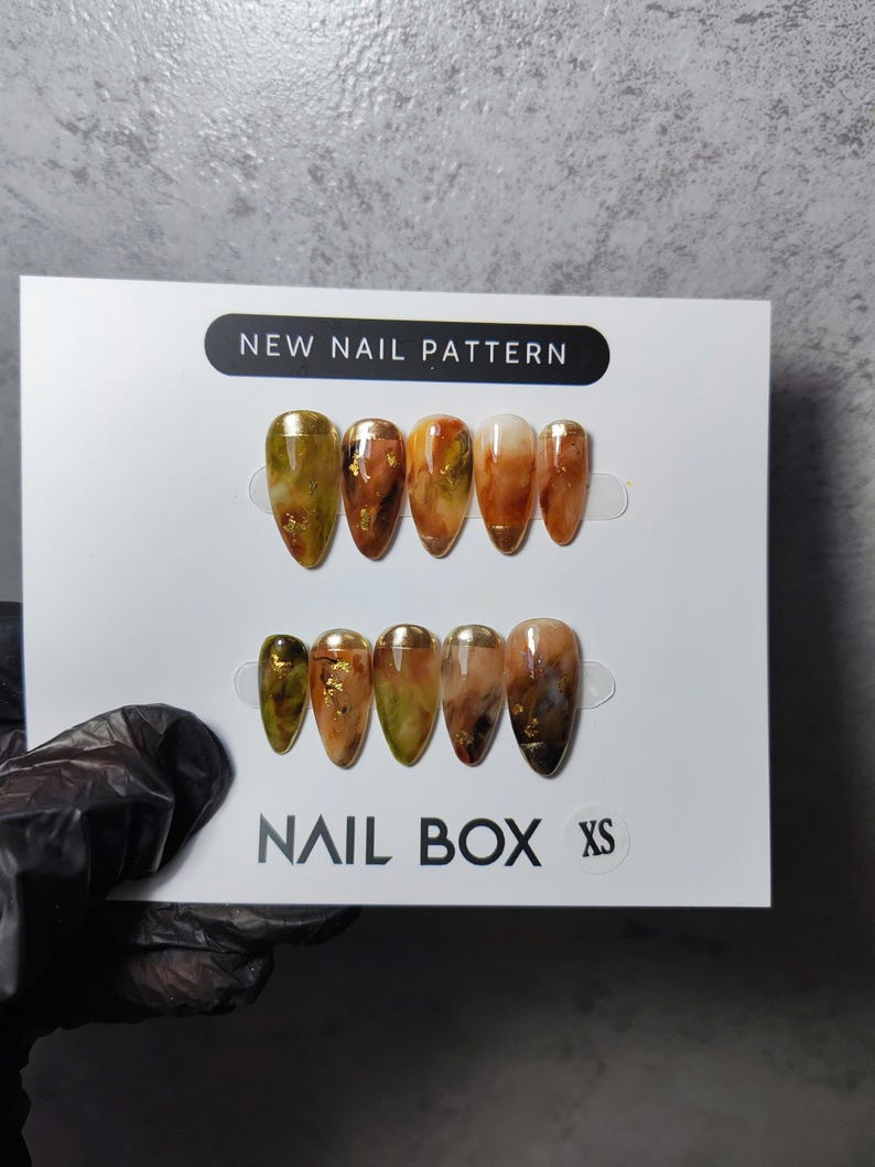 以下が含まれることがあります： Nail Box XS パッケージに入ったアーモンド型のつけ爪のセット。爪は、茶色、金、緑の色合いの大理石模様で、金箔のアクセントが付いています。パッケージには「NEW NAIL PATTERN」と「NAIL BOX」の文字があります。