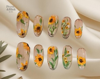May include: A set of ten clear press-on nails with a sunflower design. Each nail features a different arrangement of yellow sunflowers with green leaves and stems. The nails are oval-shaped and have a glossy finish. The text "Press & Blink by NA" is visible.