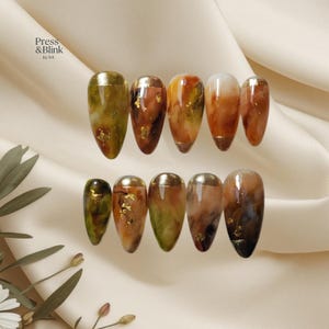 May include: A set of press-on nails with a gold metallic tip and a marbled design in shades of green, brown, and orange. Some nails feature gold leaf accents. The text "Press & Blink by NA" is visible.