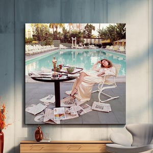May include: A large framed print depicts a woman in a cream-colored outfit lounging by a pool. An Oscar statue sits on a table surrounded by newspapers. The scene is set outdoors with palm trees and a pool in the background.