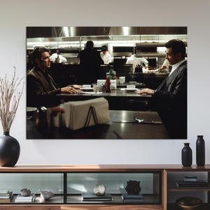 May include: A large framed print of a movie scene hangs above a modern wooden console table. The table has a glass-fronted cabinet and open shelves. A black vase with dried branches and two black bottles are on the table.