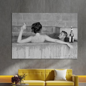 May include: Black and white wall art print of a couple in a stone tub, one smoking a cigarette, the other with a bottle. The artwork hangs above a yellow sofa with a white pillow, a small table, and a rug.