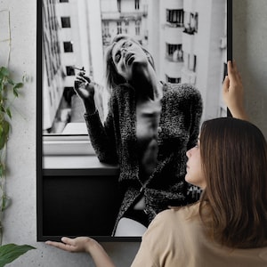 May include: Black and white framed print of a woman leaning out of a window, holding a cigarette. The woman is wearing a textured cardigan. The print is held by a person wearing a tan crop top.
