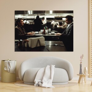 May include: A framed print of a scene from a film, featuring two men seated at a table in a restaurant. The image has a dark, cinematic aesthetic, with a focus on the characters and their interaction. The background shows a kitchen.