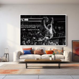 May include: Black and white framed print of a mixed martial arts fighter celebrating a victory. The image features a quote by Conor McGregor. The print hangs above a white sofa with colorful throw pillows in a modern living room setting.