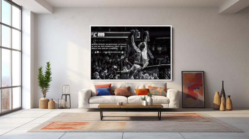 May include: Black and white framed print of a mixed martial arts fighter celebrating a victory. The image features a quote by Conor McGregor. The print hangs above a white sofa with colorful throw pillows in a modern living room setting.